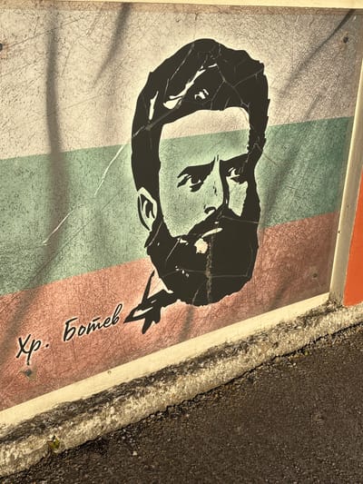 An outdoor mural featuring a stylized black and white portrait of a man with a beard and intense eyes is painted on a wall that appears to be part of the Enevo town hall in Enevo, Bulgaria. The portrait is superimposed on the Bulgarian national flag, with horizontal stripes of white, green, and red. Below the portrait, in Cyrillic script, is the name "Хр. Ботев" (Hristo Botev), a prominent Bulgarian revolutionary and poet. The mural shows signs of wear and tear, with cracks and scratches visible on the surface. The lower portion of the image shows the rough texture of the wall base and the asphalt ground in front of it. The lighting suggests it is daytime, with shadows casting diagonally across the mural.