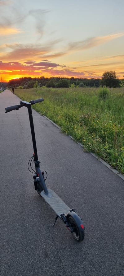 An electric scooter is parked on a paved path near a grassy field at sunset. The sky is a vibrant mix of orange, yellow, and pink hues, with streaks of clouds. A lone figure walks on the path in the distance, silhouetted against the bright sky. The path is bordered by a strip of concrete and a field of tall green grass with some scattered wildflowers. The scene is peaceful and serene, characteristic of a suburban park or recreational area. The location is identified as Romashkovo, Russia.