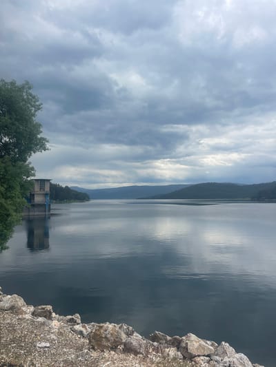 The image presents a landscape view of a lake with a cloudy sky overhead. A mountain range is visible in the distance across the lake. In the foreground, a water control structure with pipes extending into the water is present. Trees are clustered around the structure. The edge of a rocky terrain or cliff is visible on the right side of the frame. The location is identified as Sv. Konstantin, Bulgaria. There are no people depicted in the image.
