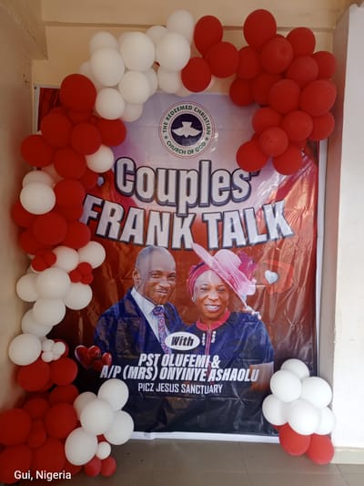 A banner for "Couples Frank Talk" is displayed, featuring portraits of a smiling man and woman. The man, wearing a suit and tie, is on the left, while the woman, in a blue dress and pink headwear, is on the right. The banner also includes text identifying "PST OLUFEMI & A/P (MRS) ONYINYE ASHAOLU" and "PICZ JESUS SANCTUARY". The banner is partially framed by a balloon arch made of red and white balloons, cascading down the left side and bunching at the bottom right. The scene is set outdoors against a textured wall, with the bottom right corner showing a tiled floor. Text at the bottom left reads "Gui, Nigeria".