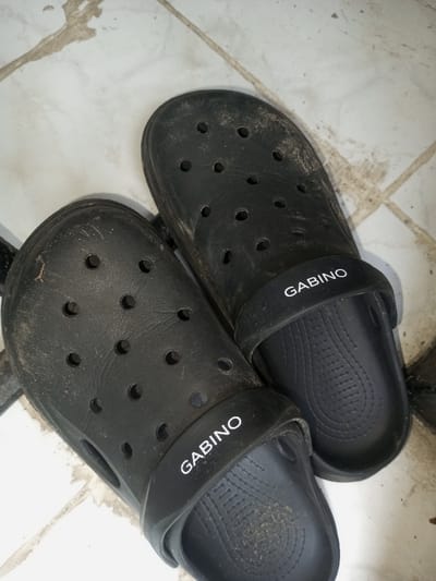 Two black, well-worn "GABINO" brand clog-style shoes rest on a dirty tile floor. The shoes are positioned close together, with one slightly overlapping the other. Both are covered in dirt and grime, especially on the upper surfaces with multiple ventilation holes. The "GABINO" logo is printed in white on the strap of each shoe. The tile floor appears to have a mix of light and dark discoloration.