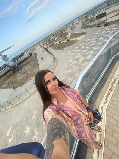 A woman with long, dark hair is taking a selfie at an outdoor park. She is wearing a pink and purple patterned shirt and pants, and an orange top underneath. She has a tattoo on her arm, and is wearing yellow nail polish. In the background, an airplane is on display, and there is a body of water visible. The park has paved walkways, benches, and trees. There are other people in the park, some sitting on benches and others walking. The sky is blue with some clouds. The location is the Arablyar city park in Russia.
