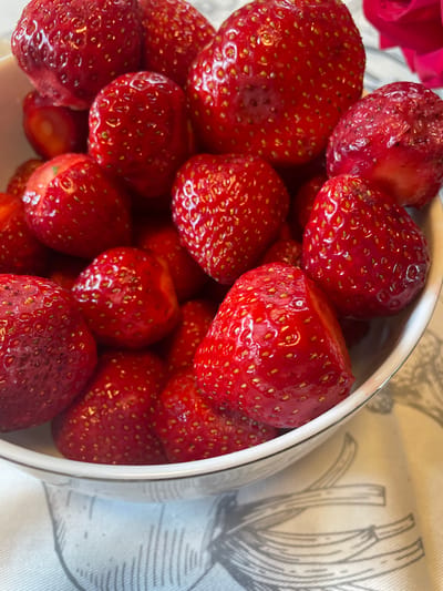 A close-up, slightly elevated shot captures a white bowl filled with plump, ripe strawberries. The strawberries are a vibrant red, with visible seeds dotting their surfaces. Some strawberries have a dewy sheen, suggesting they are freshly washed or have a natural glaze. The bowl is positioned on a white cloth with a faint gray floral or botanical pattern. A blurred pink or red rose petal is visible in the upper right corner of the image. The lighting highlights the texture and juiciness of the fruit. The location is not specified.