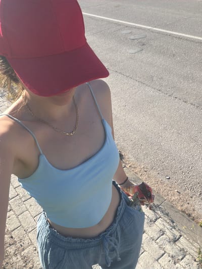A woman wearing a light blue top, grey shorts, and a red baseball cap stands on a brick and concrete sidewalk adjacent to an asphalt road. She holds a bag of chips and wears a gold chain. The location appears to be an outdoor public space in Lesichovo, Bulgaria.