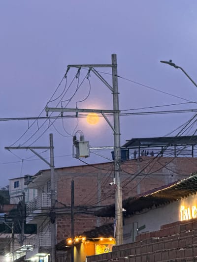 The image displays an urban twilight scene in Timaná, Colombia, viewed from a low angle. The sky, rendered in shades of purple and blue, features a large, luminous orange-yellow full moon positioned prominently in the upper mid-frame, partially obscured by light atmospheric haze. The foreground and midground are dominated by a dense network of overhead utility infrastructure.

Multiple utility poles, likely concrete or wood, rise vertically through the scene. The most prominent pole is centrally located, extending high and supporting several cross-arms from which numerous black electrical and communication wires crisscross horizontally and diagonally across the frame. A grey, rectangular electrical transformer is suspended from a cross-arm on this central pole, positioned to the left of the moon. To the far right, a black streetlight fixture extends diagonally upwards from another pole. A small, white security camera is mounted on the central utility pole, facing downwards.

In the background, various urban structures are visible. On the left, a multi-story building with white walls and an exterior white metal staircase is present, with a green hillside visible behind it. In the center, behind the main utility pole, an unfinished or exposed brick building rises. Further to the right, a building with a dark, possibly tiled or textured, roof is visible. On a central rooftop, a dark water tank and a dark, possibly corrugated metal, roof structure are present, with several small, dark shapes consistent with birds perched on a horizontal beam. On a lower building facade to the right, illuminated yellow-orange letters, partially spelling "ORA CAFE," are visible, alongside decorative string lights. No individuals are depicted, and no explicit actions or interactions are occurring within the static scene.