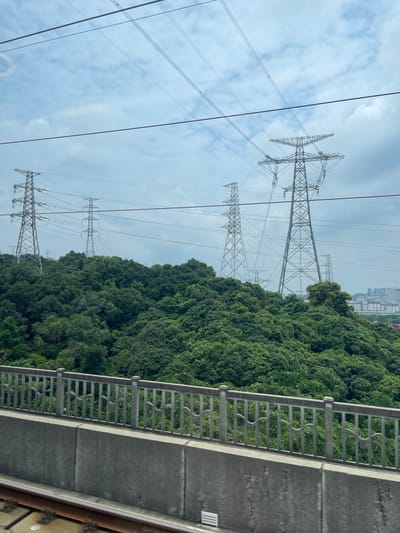 The image captures a scene looking upwards, with a focus on multiple towering electricity pylons against a cloudy sky. The pylons are made of lattice steel and support thick electrical cables that stretch across the frame. To the right of the pylons, a dense, lush green forest blankets a hillside, suggesting a natural or semi-natural setting. In the background, a cityscape with numerous buildings is visible, indicating the presence of an urban area. In the foreground, a concrete walkway with a metal railing runs parallel to the edge of the frame, implying the viewpoint is from a raised structure like a bridge or elevated road. The overall impression is one of infrastructure and nature coexisting. The time of day appears to be overcast daylight, with no strong shadows visible. The location is identified as Guangming District, China, which aligns with the presence of significant power infrastructure and a mix of natural landscape and urban development. No people are visible in the image.