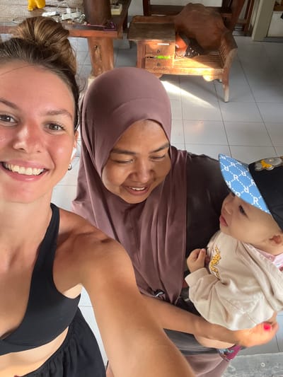 A selfie-style, indoor photograph captures three individuals in Pemenang Timur, Indonesia. In the foreground, a young woman with light brown hair pulled into a bun smiles broadly at the camera. She is wearing a black athletic top and black shorts. To her right, a woman wearing a mauve hijab and a dark top is looking down and smiling. She is holding a baby, who is wearing a light-colored hoodie and a blue and white patterned cap. The baby is looking towards the woman in the hijab. In the background, a wooden table with drawers and a carved wooden chair are visible. The floor is tiled. The overall lighting suggests daylight.