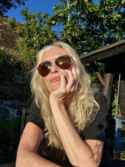 This is a close-up, angled outdoor photograph of a woman wearing large, reflective sunglasses. She has long, blonde hair and her face is partially obscured by her hand. She is wearing a black top. The background is filled with lush green foliage, a large tree with leaves, and a bright blue sky, indicating it is daytime and the weather is sunny and clear. The image has a relaxed, perhaps contemplative feel. The woman appears to be enjoying a sunny day outdoors. There is no visible text.