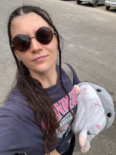 A woman wearing sunglasses and a blue T-shirt lies on a concrete surface. A stuffed animal resembling a hamster rests on her chest. The location is not specified.
