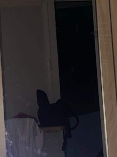 The image shows a dark interior, possibly a room or a storage area, with a large dark rectangle dominating the center, which appears to be a screen or a window reflecting light from the outside. On the right side of the dark rectangle, a dark, amorphous object is visible, which could be a piece of furniture or a bag. To the right of this object, a light-colored, translucent bag or bundle is partially visible, with some reddish material peeking out from underneath. The overall lighting is dim, suggesting an indoor environment with limited natural light. The scene appears static, with no clear activity taking place. No text is visible in the image.