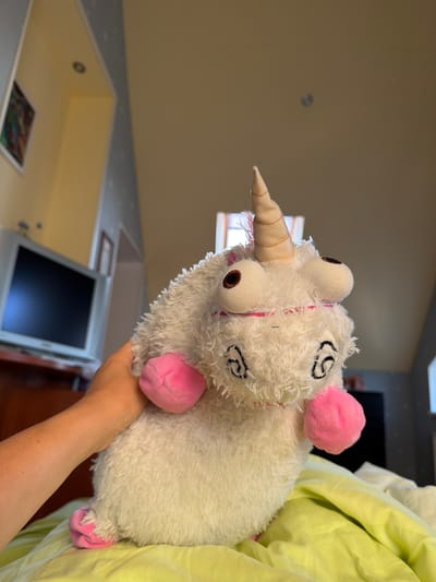 A white, fluffy plush toy unicorn with pink hooves and a beige horn is held in a person's hand. The unicorn has large, round, brown eyes with black pupils and stitched eyebrows. A thin pink line runs across its face, resembling a mouth. The toy is resting on a bright yellow-green sheet. In the background, a television set is visible on a dark wooden dresser. The room appears to be a bedroom with a sloped ceiling and a window. The setting is likely a residential area.