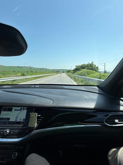 The image is taken from inside a car, showing the driver's perspective of a road. The weather is sunny with a clear blue sky. On the left, there is a green field and trees along the road. In the distance, another car can be seen driving on the road. The dashboard of the car is visible, with a GPS screen displaying a map with the words "31 KM", "14 и 9 мин" and the number 65. Additionally, the GPS screen indicates that the location is "ул. Акация Благуна, Гоца." A brand name "Areon" is visible near the car's air vents. The image doesn't have any readily discernible indication of a city or region; however, it says ул. Акация Благуна, Гоца.