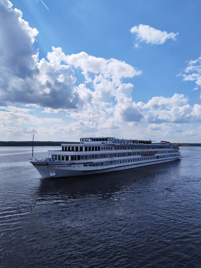 The image depicts a white, multi-deck cruise ship, named "Nikolai Chernyshevsky" in Cyrillic script on its hull, sailing on a dark blue body of water. The ship is oriented towards the left of the frame. In the background, a thin strip of land with dark green trees is visible. The sky is bright blue with large, fluffy white clouds and contrails of an airplane. The water shows subtle ripples and reflections of the sky. The location is most likely Pruzhinino, Russia, given the Cyrillic writing on the ship.