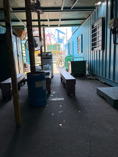 The image depicts the interior of a shipping container converted into a workshop or storage space, likely in Mokpo-si, South Korea. The container walls are a corrugated blue metal. The scene is cluttered with various objects, suggesting ongoing work or storage of materials.
In the foreground, a yellow metal ladder structure is visible, with hazard tape on its horizontal supports. Two black oscillating fans are positioned on the ladder. A crumpled paper bag hangs from one of the supports. Further back, a large blue cylindrical container is prominent, with visible text reading "UNI-TECH" and other labels. Next to it, a white air conditioning unit is visible.
The scene is illuminated by natural light filtering in from the open end of the container, which reveals construction or industrial activity outside, including cranes and buildings. The floor is a dark, rough asphalt or concrete surface.
Notable details include the presence of industrial equipment like fans and the large drum, indicating a functional space rather than a living area. The overall impression is one of a busy, perhaps temporary, workspace. The time of day appears to be daytime, with ample natural light. There are no people visible in the frame. The text on the blue drum provides a brand name and potentially product information. The location context of Mokpo-si, South Korea, suggests an industrial or port area.