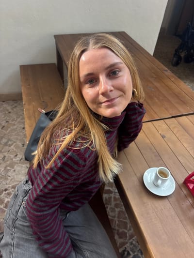 The image displays a female subject seated at a wooden table within an indoor establishment, identified as being in Porto Salvo, Portugal. The female has long, straight blonde hair with a middle part, fair skin, and blue eyes. She is wearing a long-sleeved, high-necked sweater with alternating horizontal stripes of dark red and dark grey, paired with grey jeans. She is looking directly at the camera with a subtle smile, with her right arm resting on the table surface. To her right, on the wooden tabletop, a white ceramic espresso cup holding dark liquid is positioned on a white saucer. A dark-colored handbag with a visible brown strap accent is situated on the wooden bench to her left. The furniture comprises robust wooden tables and integrated benches. The flooring consists of small, irregularly shaped reddish-brown and dark grey tiles. The background features light-colored walls, and a dark, indistinct object, possibly a wheeled item such as a stroller, is partially visible in the distant right section of the frame.