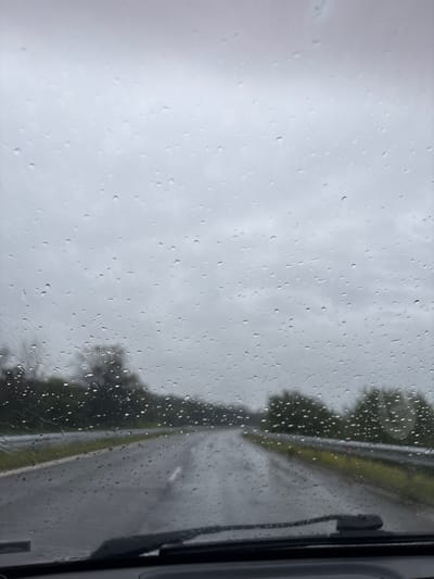 The image depicts a view from inside a vehicle, looking out through a rain-splattered window. The entire visible surface of the window is covered in numerous water droplets of various sizes, indicating active rainfall.
The scene outside the window is heavily blurred due to the rain and possibly motion or shallow depth of field, but general elements are discernible. A grey, overcast sky occupies the upper-left portion of the frame, suggesting a dull, possibly afternoon or morning, time of day. On the right side, blurred dark green foliage, likely trees, lines what appears to be a wet road or highway. The road surface reflects light, confirming its wet condition.
On the far right edge of the image, parts of the vehicle's interior or exterior structure are visible, including what seems to be a dark-colored side mirror or the frame of the window, and a portion of a windshield wiper blade. This detail confirms the perspective is from within a car or similar vehicle.
No specific activity is clearly captured beyond the general experience of being in a vehicle during rain. There are no people visible. The location context is Kazanluk, Bulgaria, suggesting this rainy scene is from that region. There is no visible text in the image.