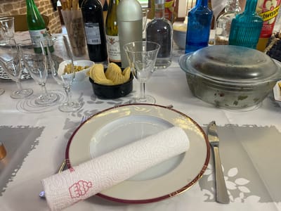 This image depicts a dining table set indoors in Ancenis, France. The table is covered with a white tablecloth overlaid with a light grey fabric featuring a subtle floral or leafy pattern. In the foreground, a white dinner plate with a gold and deep red decorative rim holds a roll of white paper towel, embossed with a pink stylized cake or cupcake illustration. To the right of the plate, a silver-colored butter knife is positioned.
Further back, several empty clear glass champagne flutes and wine glasses are arranged on the left side of the table. A black bowl filled with golden potato chips (crisps) and a small white bowl containing green olives are visible in the mid-ground. A large clear glass bowl with a matching clear glass lid, containing a green and reddish-brown mixture consistent with a salad, occupies the right side of the frame.
Assorted bottles are distributed across the table: a green glass bottle labeled "BIO," a dark glass bottle identified as "MANUS DOMINI" red wine, a light-colored glass bottle of "Domaine ROAUX" white wine, a clear glass bottle with dark liquid (likely spirits), two vibrant blue glass bottles, and a yellow-labeled "Schweppes Pomme" bottle. A paper bag containing breadsticks is also visible behind the chips and wine bottles. The background is blurred, showing light-colored walls, possibly brick or stone, indicating an interior setting. No individuals are present in the scene.