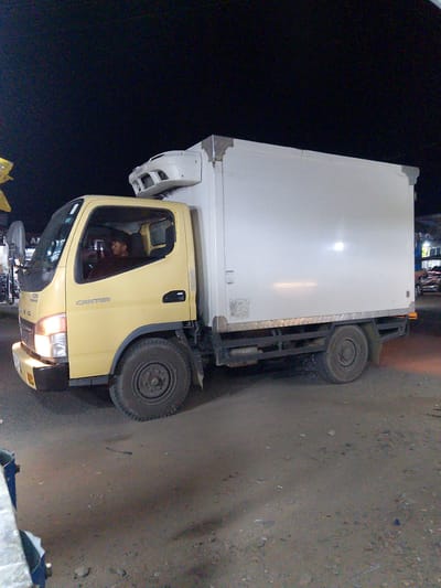 A yellow truck with a white refrigerated box body is parked on a dirt road at night. The truck has the word "CANTER" written on its side. A man is visible inside the driver's cabin. The background is dark, indicating it is nighttime, and there are some lights and buildings visible in the distance, suggesting an urban or semi-urban environment. A portion of a blue metal structure is visible in the foreground on the left side of the image. The ground appears to be unpaved and uneven, with some debris scattered on it. The scene is located in Gampongpaya, Indonesia.