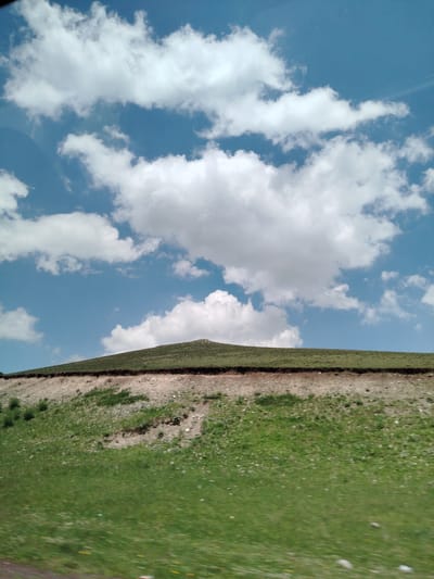 A wide-angle, low-angle outdoor photograph captures a bright blue sky filled with fluffy white clouds. The sky occupies the upper half of the frame. Below the sky, a gently sloping green hill dominates the lower half of the image. The hill's surface is covered in short, vibrant green grass, dotted with occasional lighter green patches and small yellow flowers. A distinct horizontal line of dark brown soil separates the grassy upper slope from a lighter, rockier embankment below. This embankment appears to be eroded, with scattered stones and sparse patches of green vegetation. The ground in the foreground is a blur of motion, indicating the photograph was taken from a moving vehicle. The scene is devoid of any people or man-made structures, suggesting a rural or natural setting, possibly on the outskirts of a town or village. The overall impression is one of natural beauty and open space.