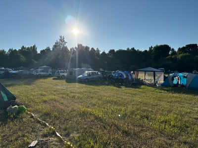 The image depicts a campground in the Валожынскі сельскі Савет, Belarus, under a bright, sunny sky with visible lens flare from the sun. The scene features a field of grass with several tents and parked cars. In the foreground, a green tent is partially visible on the left, along with some bags and yellow caution tape on the ground. Further into the field, there are various tents, including a large blue tent on the right side of the image and a white gazebo-like structure. Several cars are parked among the tents, including a silver hatchback and a dark sedan. In the background, a dense line of trees borders the campground. There are no people clearly visible in the frame, but there are motorcycles parked near some of the tents. The overall impression is that of an outdoor event or festival at a campsite.