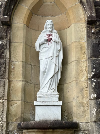 This media file depicts a statue of Jesus Christ, shown from the chest up, carved in white marble. The statue is set within a stone alcove, which is part of an exterior wall of a building. The alcove is constructed with blocks of weathered, light-colored stone, forming curved and straight surfaces. Above the alcove, rough-hewn, dark stone blocks form the upper part of the wall, with some patches of lichen or moss visible. Below the statue, a base is visible, also made of white marble with some grey veining. The statue is positioned on this base. Jesus is depicted with flowing hair and a beard, and his face appears serene. His right hand is raised, touching his chest where a stylized, red heart is prominently displayed. Rays emanate from the heart, suggesting a sacred representation. The overall impression is one of religious devotion and artistry. The lighting suggests daytime, and there are no immediate cues about specific weather conditions, though the lichen on the stone implies a generally humid or temperate climate. The stone construction and style of the statue might suggest a historical or ecclesiastical building. The location context of Mountcharles, Ireland, further suggests this is likely an outdoor religious monument or part of a church or cemetery. No visible text is present in the image.