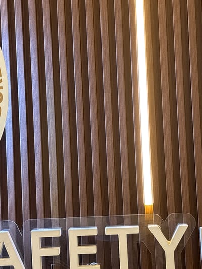 The image presents an interior scene, likely a commercial or public space, featuring a wall covered in horizontal, dark brown wooden slats. A bright, white LED light strip runs horizontally across the upper middle section of the wall, casting a clear glow and highlighting the textured grain of the slats.
On the right side of the image, a vertical sign is prominently displayed. It consists of large, white, three-dimensional letters mounted on a clear acrylic panel, creating a slight stand-off from the slatted wall. The visible letters of this sign, read from top to bottom, are "Y", "T", and "E". Below the "E", the top curve of another letter, possibly an "O" or "C", is partially visible.
In the bottom left corner of the image, a portion of a separate sign is visible. This sign appears to be light-colored with dark lettering, and the visible text consists of the letters "ON".
No people are present, and no specific activity or event is taking place; the image captures a static view of the architectural elements and signage within the space. The artificial lighting suggests an indoor setting where the time of day is not apparent.