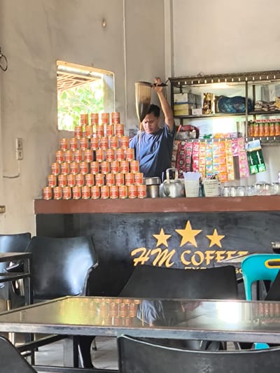 In Meunasahtutong, Indonesia, a man is preparing a drink at a coffee stand. The stand is decorated with three yellow stars above the words "HM COFFEE". Stacks of canned goods, likely food or beverages, are piled high against a wall to the left of the counter. The man, wearing a blue t-shirt, is holding a metal strainer over a cup, pouring liquid from it. Behind the counter, shelves are stocked with various items, including bottled drinks and packaged goods. Several black chairs are arranged around tables in front of the counter. A small, bright blue stool is visible to the right.