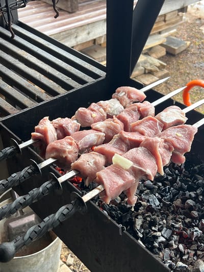 The image shows metal skewers loaded with raw meat chunks, likely pork or beef, resting over a charcoal grill. The coals beneath the meat are hot and glowing. The skewers have twisted, dark-colored handles. The grill is a black metal construction. The location is unknown.