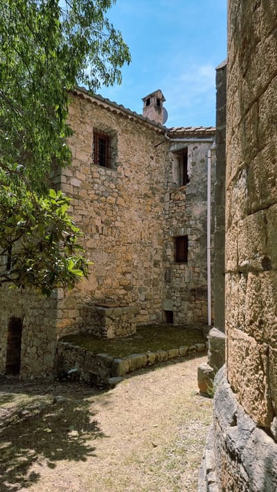 This image depicts a view of a rustic, stone-built structure with a tiled roof under a clear blue sky, likely in the town of Sales de Llierca, Spain. The foreground is a dry, grassy area with scattered pebbles and a prominent shadow cast by a tree on the left. The main structure, built from irregular-shaped stones, features a small window with a metal grate and a brick chimney protruding from the roof. Adjacent to this building, another stone wall, with much larger, more rough-hewn stones, forms a corner on the right side of the frame, suggesting an older or more substantial construction. A white pipe runs vertically down the side of the building. There are no people visible in the scene. The overall impression is one of an old, possibly abandoned, rural building.