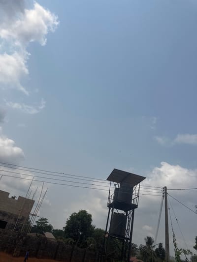 The image depicts an outdoor scene in Agbogugu, Nigeria, under a bright, partly cloudy sky. The dominant features include a tall, dark-colored metal water tower on the right, equipped with two vertically stacked cylindrical water tanks and topped with a flat, dark protective roof. To the immediate right of the water tower stands a utility pole supporting multiple horizontal power lines, on which two small birds are perched. Small, dark specks, possibly birds, are visible in the sky above the water tower.
In the mid-ground on the left, an unfinished concrete block structure with visible bamboo scaffolding indicates ongoing construction. Below this structure, a dark, low-rise perimeter wall extends horizontally across the frame. Behind the wall and water tower, various green trees and foliage are present, including identifiable palm trees. The foreground consists of reddish-brown earth. A lone individual, dressed in a blue shirt and dark trousers, is partially visible walking from left to right in the lower-left corner, mostly obscured by the perimeter wall. The scene suggests a semi-urban or rural environment with developing infrastructure.
