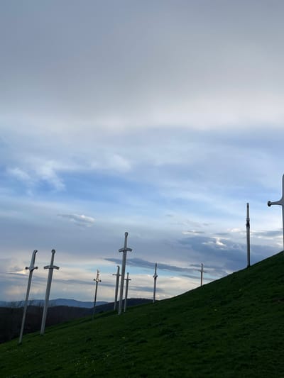 The image shows a monument of several swords planted into a grassy hillside. The background consists of a cloudy sky and a distant treeline. The location is likely Zemo Nichbisi, Georgia. There are no people in the shot.