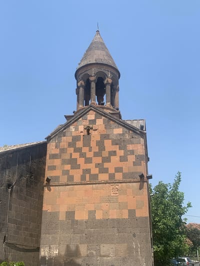 The image shows a low-angle, full shot of the bell tower and adjoining structure of the Saghmosavank Monastery, located in the city of Karbi, Armenia. The sky is clear and blue. The bell tower, made of dark stone, has a conical roof and is supported by columns. The main structure of the monastery features a checkerboard pattern of light brown and dark grey stones. A carved inscription and a decorative relief are visible on the stone facade. Several modern floodlights are attached to the exterior walls. A lush green tree grows to the right of the building. The left side of the monastery structure is made of dark, rough-hewn stone.