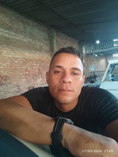 This image depicts a single adult male situated within what appears to be a workshop or garage environment in Ureña, Venezuela. The male, appearing to be of Latin American descent, is positioned in the central foreground, looking directly at the camera with a neutral expression. He has short, dark hair and is wearing a black short-sleeved t-shirt. A black digital or analog watch with a black strap is present on his left wrist. His left forearm rests on a light-colored surface, possibly a component of a vehicle. The background to the left consists of a weathered brick wall with visible mortar and faint reddish markings. To the right, a white vehicle, possibly a truck or SUV, is partially visible with its engine compartment area exposed. Overhead, a dark, corrugated metal roof supported by concrete pillars or beams is evident. The lighting suggests an indoor or semi-enclosed setting. A timestamp in the bottom right corner indicates "17/03/2026 17:49".