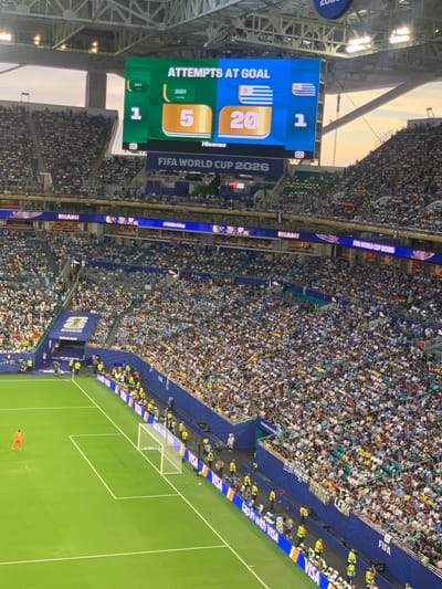 This image captures a wide-angle view of a packed soccer stadium in Miami Gardens, United States of America, likely during a FIFA World Cup 2026 event. The stadium is filled with a dense crowd of spectators wearing a variety of clothing, predominantly in light colors. A large digital scoreboard is prominently displayed, showing "ATTEMPTS AT GOAL" with statistics for two teams, indicated by their flags and numbers: one team has 1 attempt and the other has 5 attempts. Below the scoreboard, the text "FIFA WORLD CUP 2026" is visible. The stadium features tiered seating that extends high above the green soccer field. On the field, a single person in an orange uniform, possibly a goalkeeper, stands near a goal. The perimeter of the field is lined with advertisements, including those for "VISA" and "MIAMI." The scene is illuminated by natural light, suggesting it might be late afternoon or early evening, with the sky visible at the top of the image. The overall atmosphere is one of a large-scale sporting event.