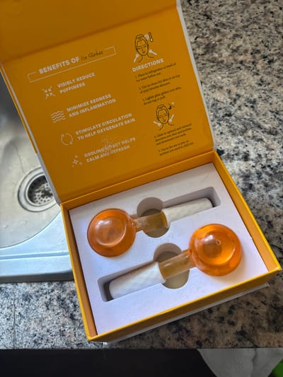 An overhead shot captures an open, rectangular yellow box containing two facial "Ice Globes" positioned on a speckled gray kitchen or bathroom countertop, with a stainless steel sink partially visible in the bottom left. The box features white molded inserts that cradle the two globes. Each globe has an orange, spherical top, a clear, light-orange stem, and a white, textured handle.
The inside lid of the box provides text about the product. On the left side, under the heading "BENEFITS OF Ice Globes," points include: "VISIBLY REDUCE PUFFINESS," "MINIMIZE REDNESS AND INFLAMMATION," "STIMULATE CIRCULATION TO HELP OXYGENATE SKIN," and "COOLING EFFECT HELPS CALM AND REFRESH." The right side of the lid, labeled "DIRECTIONS," outlines usage steps: "1. Place in refrigerator or bowl of ice water before use. 2. Use on clean dry skin or on top of your favorite skincare. 3. Lightly glide globes over skin, do not tug or pull. 4. Glide in upward and outward direction over face and jawline, and downward over neck. 5. Use in the am or pm, or anytime you need to chill out." The overall scene suggests a product ready for use in a personal care routine.