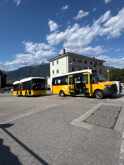 In Magadino, Switzerland, two yellow buses are parked in an outdoor lot with a light-colored, multi-story building and mountains in the background. The building, labeled "CASA COMUNALE," has multiple windows and awnings. The bus in the foreground is a Mercedes-Benz Sprinter, featuring an open passenger door and a driver visible inside. The bus behind it is a standard bus with its doors closed. The ground is paved with asphalt and a section of brickwork with a metal grate. The sky is blue with scattered white clouds. The overall scene depicts a sunny day in a semi-urban environment with public transportation.