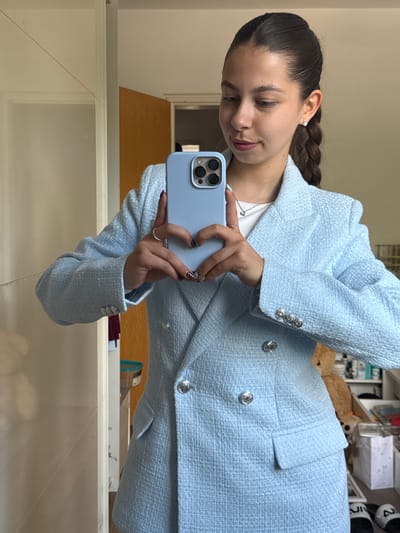 A young woman is taking a selfie in a mirror. She is wearing a light blue tweed blazer and skirt with silver buttons. Her dark hair is pulled back in a braid. She holds a light blue iPhone in her hands. The background appears to be a room with shelves and various objects, but is blurry. The location is Bruchköbel, Germany.