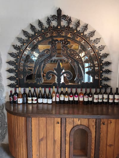 In Voskevaz, Armenia, a wooden cabinet with a rounded front is topped with a dark wooden surface displaying a collection of 18 wine bottles. The bottles are arranged in a line, with labels facing forward, and vary in color, including red, white, and rosé wines. Behind the bottles, a large, ornate black metal mirror with decorative cutouts frames a reflection of a brick wall and what appears to be a wooden table and chairs, possibly in a winery tasting room or restaurant. The mirror's design features two stylized bird-like figures facing each other, outlined in a copper or bronze color. The overall impression is of a display within a wine-related establishment.