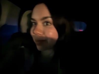 The image depicts a dark, motion-blurred interior scene within a vehicle at night. A single individual, appearing to be a young person with dark hair and fair skin, is prominently positioned on the right side of the frame, facing towards the left. Their eyes are wide open, conveying an alert or surprised expression. They are dressed in a dark-colored upper garment. The background indicates a vehicle interior, with vague shapes suggesting seats or headrests. In the far right background, a blurred rectangular reflection contains a horizontal red light streak, indicative of a vehicle's taillight viewed through a mirror while in motion. No specific landmarks or distinguishing features are present to identify a precise location within Veracruz, Mexico.