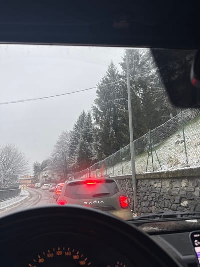 The image displays a traffic scene in Lipomo, Italy, during winter conditions, viewed from inside a vehicle. The foreground reveals the top of a steering wheel and dashboard. The road ahead is occupied by a line of stationary or slowly moving vehicles. Immediately ahead is a dark green Dacia SUV, identifiable by its rear emblem, with its red brake lights illuminated. The road surface exhibits patches of snow and appears wet. To the right, a low, dark grey stone retaining wall runs parallel to the road, topped by a green chain-link fence. Behind the fence, a snow-dusted incline leads to a dense row of snow-covered evergreen trees. A utility pole stands near the road edge on the right. On the left side of the road, bare deciduous trees and glimpses of residential structures are visible, also lightly dusted with snow. The sky is uniformly overcast.