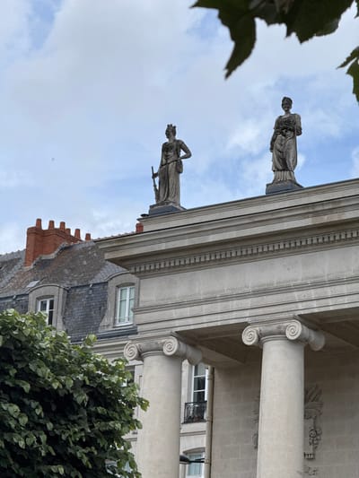 The image shows the façade of a neoclassical building with two statues of women standing on the roofline, against a cloudy sky. The building features large columns with Ionic capitals. To the left of the neoclassical building, a residential building with a steeply pitched, grey slate roof and red brick chimneys is visible. A large tree with lush green leaves is in the foreground on the left. The scene is located in Nantes, France.