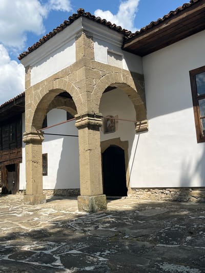 The image depicts an exterior view of a building, likely a church or a historical house, in Zheravna, Bulgaria. The architecture features traditional Bulgarian Revival style elements, including a white plastered facade, a tiled roof with wooden eaves, and stone arches supported by thick pillars. A dark wooden door is visible beneath the arches, and a small religious icon is mounted on the wall above it. The ground in front of the building is paved with large, irregular stone slabs, and sunlight casts shadows across the scene, indicating a daytime setting. The sky above is blue with scattered white clouds. To the left, a portion of an adjacent dark wooden structure is visible. The overall impression is one of a historic and well-preserved structure within a cultural heritage site.