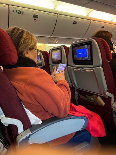 The image shows the interior of an airplane cabin, likely in flight. A blonde woman is reclined in her seat, wearing a burnt orange fleece jacket and black undershirt, and looking at her smartphone. There are screens on the back of the seats in front of her. A red blanket and blue pillow are visible. The aircraft is potentially landing in Pudahuel, Chile.