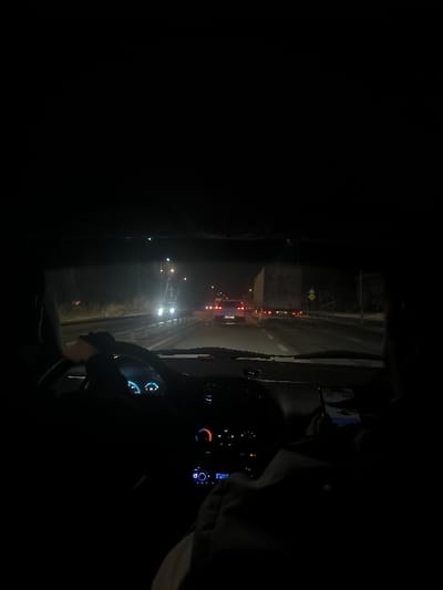 A dark car interior shows a driver at night. Headlights of cars ahead illuminate the road. The dashboard glows, with a hand on the steering wheel. A passenger to the right is looking at a phone. Traffic lines the highway, marked by illuminated signs and reflective barriers. The overall ambiance suggests a late-night drive.