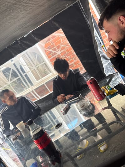 The image depicts three males gathered around a clear glass table beneath a dark canopy or tarp in Boston, United Kingdom.

On the left, an adult male with short dark hair and stubble wears a dark tracksuit top with white stripes. He is looking towards the center of the table. In front of him rests a white ceramic cup.

At the far side of the table, a male with dark, curly hair is seated, focusing on objects directly in front of him. He wears a dark long-sleeved top and appears to be manipulating a small, dark object and a white rectangular item, possibly rolling papers, on the table. Adjacent to his hands is an open pack of red and white cigarettes and a clear glass ashtray containing cigarette butts and ash.

To the right, a third male is shown in profile, looking down at the table. He has dark hair with a faded haircut and a short beard, wearing a dark long-sleeved top. His right hand is near his chin. On the table near him are a red Reign Total Body Fuel energy drink can (Peach Fizz flavor), a yellow and blue metal tin, and a black smartphone.

In the foreground, a partially consumed black Coca-Cola Zero Sugar bottle is visible, with an expiration date of "30-05-25" on its cap. Under the table, the rightmost male's feet are visible, clad in white socks and yellow slide sandals with blue stripes.

The background reveals a brick building with white-framed arched windows, indicating an outdoor or semi-enclosed patio setting. The overhead dark fabric tarp or canopy is supported by light-colored poles, with gaps allowing daylight to penetrate. The ground beneath the table appears to be concrete.