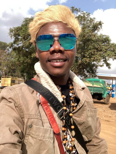 A person poses outdoors wearing sunglasses, a tan jacket with a sherpa collar, and a beaded necklace. The person has short, blonde hair and is wearing a black shirt. A black strap with white lettering crosses the jacket. In the background, trees and a green vehicle can be seen. The sky is partially cloudy.