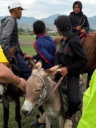 In Shabdan, Kyrgyzstan, under a cloudy sky and rolling hills, several young individuals are interacting with donkeys. In the foreground, a light brown donkey with its eyes closed is being petted by an adult's hand wearing a silver bracelet. The donkey has a colorful woven halter. To its left, another donkey, darker in color, is partially visible, with a young person in a gray hooded vest and white baseball cap seated on it. Further back, a young person in a black puffer jacket with a hood up is riding a brown horse, holding a riding crop. Another young person in a blue tracksuit with red stripes is seated on a donkey behind the first. A fourth young person, wearing a black Nike jacket and cap, is also seated on a donkey, holding a white object. The environment appears to be a grassy field with a village and distant mountains in the background.