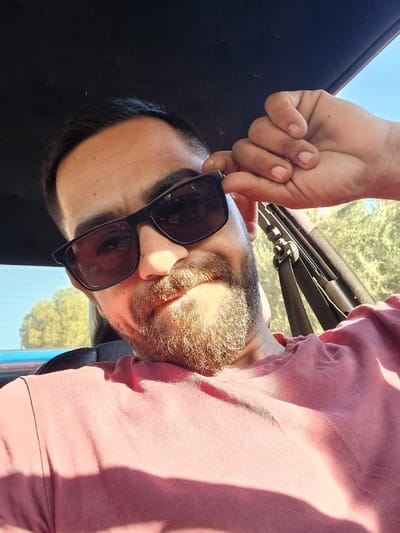 A man wearing sunglasses and a pink t-shirt is sitting in a car, with his hand resting on his forehead. The image was taken outdoors, with a clear blue sky and trees visible in the background. The sun is shining, casting shadows on the man's face and shirt. The scene is set in a car, likely in a rural or suburban area, with vegetation visible outside the window. The specific location in the Chersonisos Municipal Unit, Greece, is not discernible from the image itself.