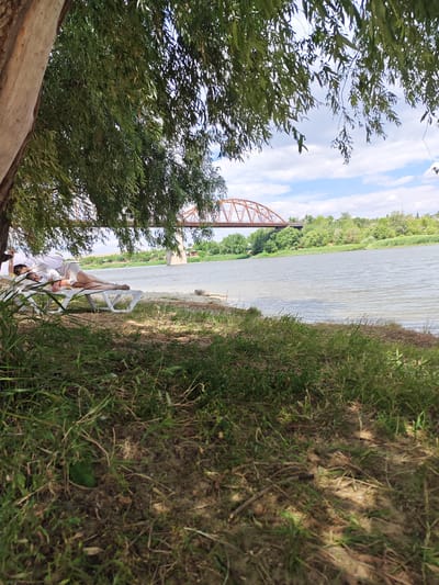 A couple is relaxing on lounge chairs by a river. The woman is lying on her side, wearing a white top and shorts, with her head resting on her arm. The man is also lying down, wearing a white shirt and shorts, with his head on a pillow. They are both under the shade of a large tree with green leaves.

In the background, a river flows gently, with a bridge spanning across it. The bridge is made of red metal and has a distinct arch design. Lush green trees line the riverbanks, and the sky is partly cloudy with patches of blue. The scene appears to be set during a warm, sunny day, likely in the afternoon, given the shadows cast by the tree. The overall mood is peaceful and relaxing, suggesting a leisurely day by the water.

The location is Belaya Kalitva, Russia. There is no visible text in the image.