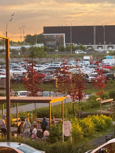 This media file depicts an outdoor scene in Drozhzhino, Russia, likely during the golden hour of sunset. The sky is filled with warm, hazy orange and yellow tones, suggesting either a clear but late afternoon or evening sky, or possibly a light atmospheric haze.
The main focus of the image is a large parking lot filled with rows of parked cars, predominantly white and silver. The cars are lined up neatly, indicating a structured parking area, possibly for a residential complex or a commercial establishment.
In the foreground and mid-ground, there are several young people engaged in various activities. Some appear to be socializing, standing in small groups and interacting. Others are seated on a low wooden structure that resembles benches or a low wall, also seemingly engaged in conversation or relaxation. One individual is leaning against a tree. The overall atmosphere among the people seems relaxed and casual.
Several trees with vibrant red foliage are scattered throughout the scene, adding pops of color against the backdrop of cars and greenery. These trees are in full leaf, suggesting a warmer season like late spring or summer. There are also streetlights visible, indicating the presence of infrastructure for illumination.
A building with dark, reflective windows is visible in the background on the left, adding a sense of urban or suburban development to the setting. On the right side, a white car is partially visible, and a white oval sign with some printed text is mounted on a pole, though the text is illegible.
The lighting and the relaxed demeanor of the individuals suggest a peaceful evening setting in a semi-urban environment.